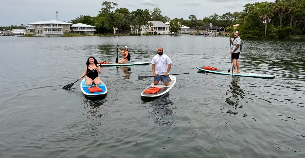 Four people paddleboarding on a calm lake with houses and trees in the background.