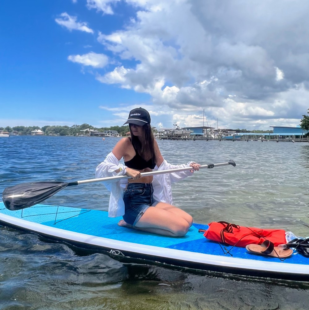 Woman in hat sitting on a paddleboard with paddle on a calm lake under partly cloudy sky.